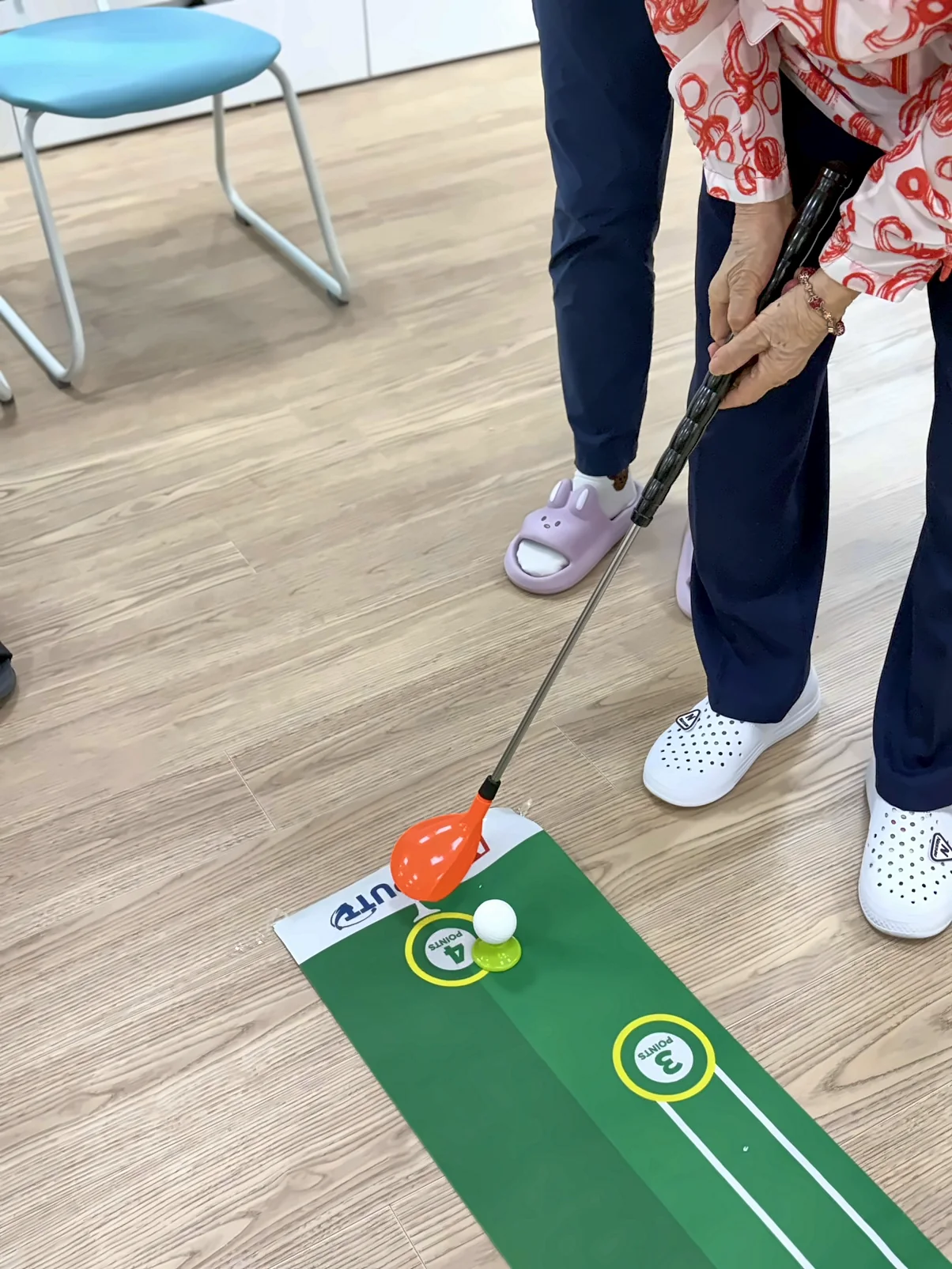 퍼팅 매트 위에서 골프채로 공을 굴리는 모습. 선생님이 곁에 서 있습니다