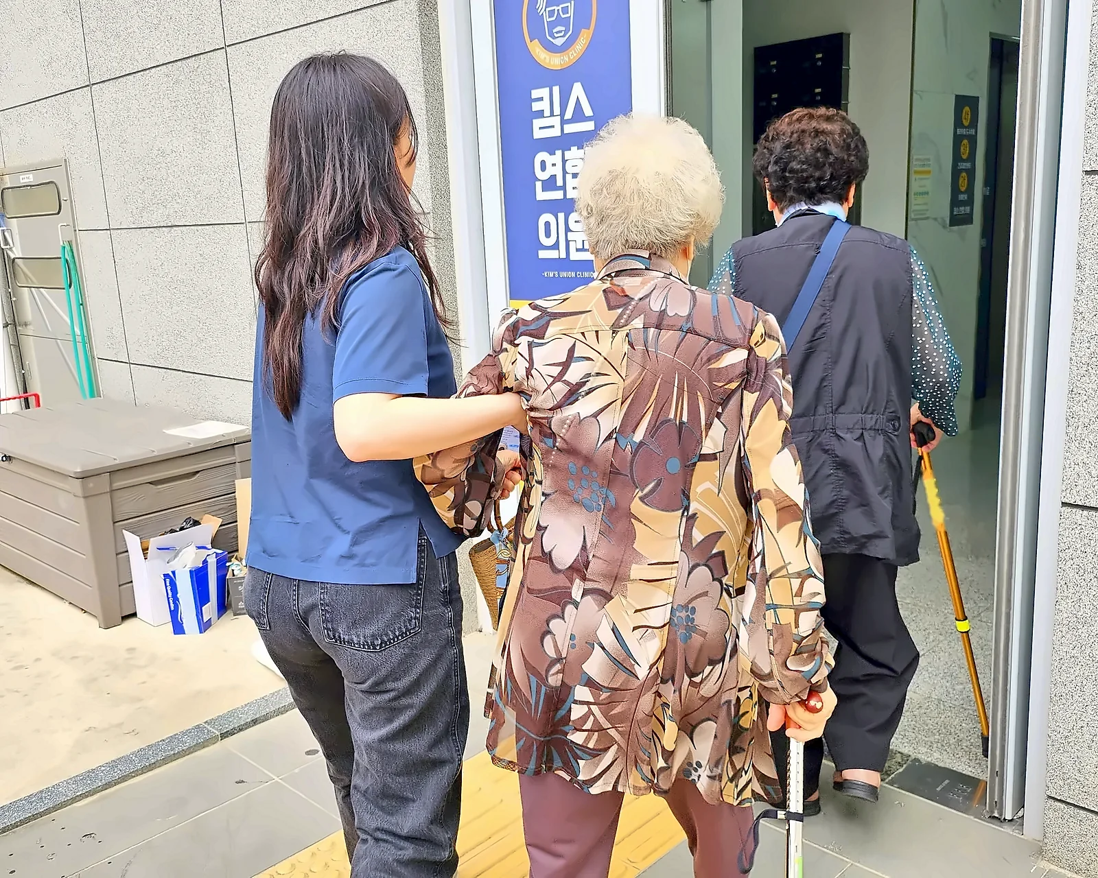 선생님이 어르신의 팔을 잡고 함께 의원 건물로 들어가는 병원 동행 뒷모습