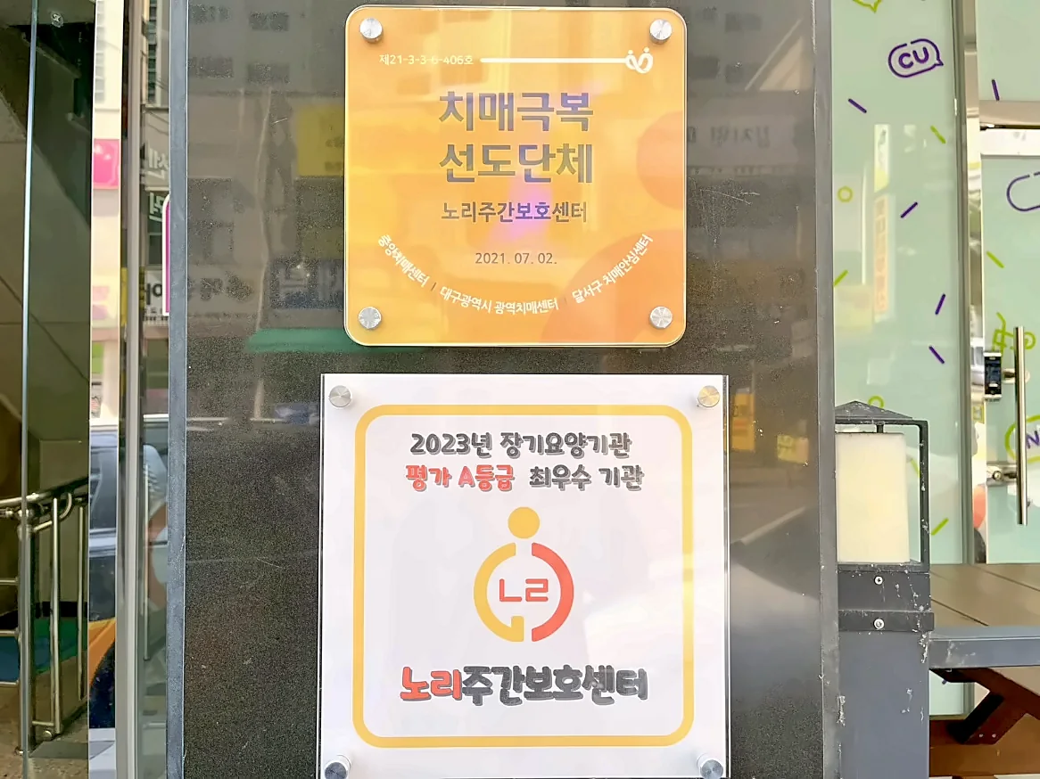센터 입구 기둥에 붙은 현판 두 개. 위는 2021년 7월 2일 치매극복 선도단체 현판, 아래는 2023년 장기요양기관 평가 A등급 최우수 기관 현판